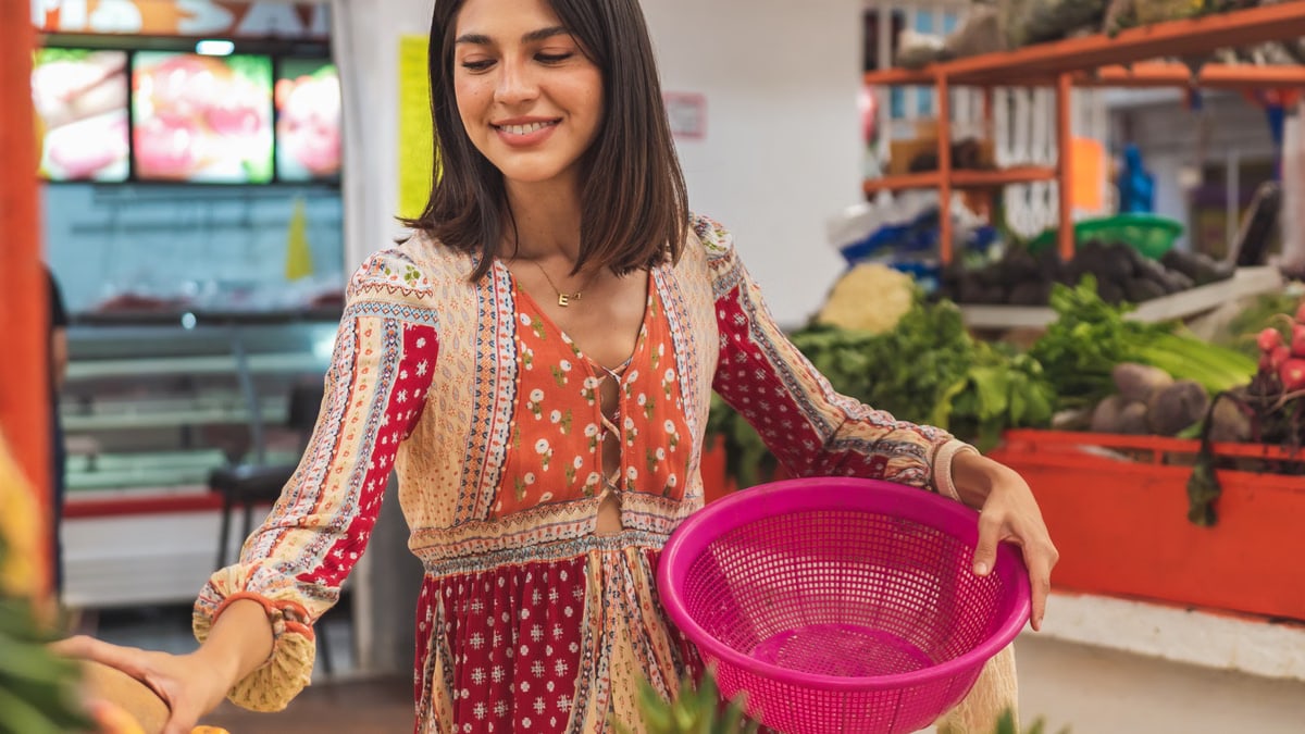 Smiling woman with a pink basket selecting fresh produce at a local market.